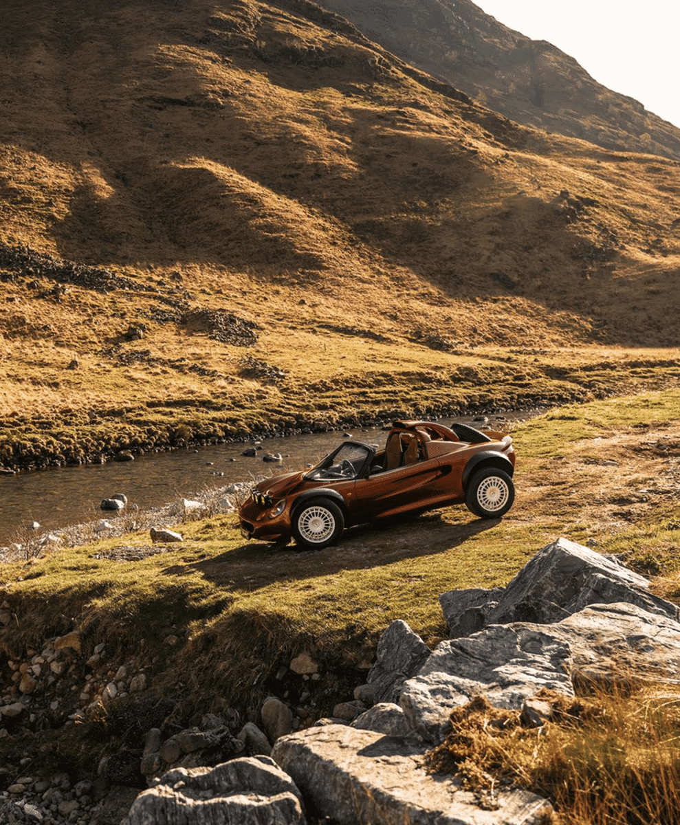 An orange convertible off-road vehicle parked in a shallow stream within a mountainous valley.