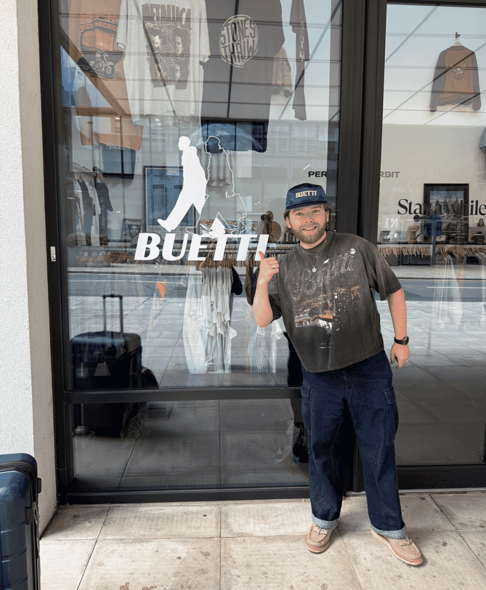 A man in a graphic tee and blue cap giving a thumbs-up outside a shop window.