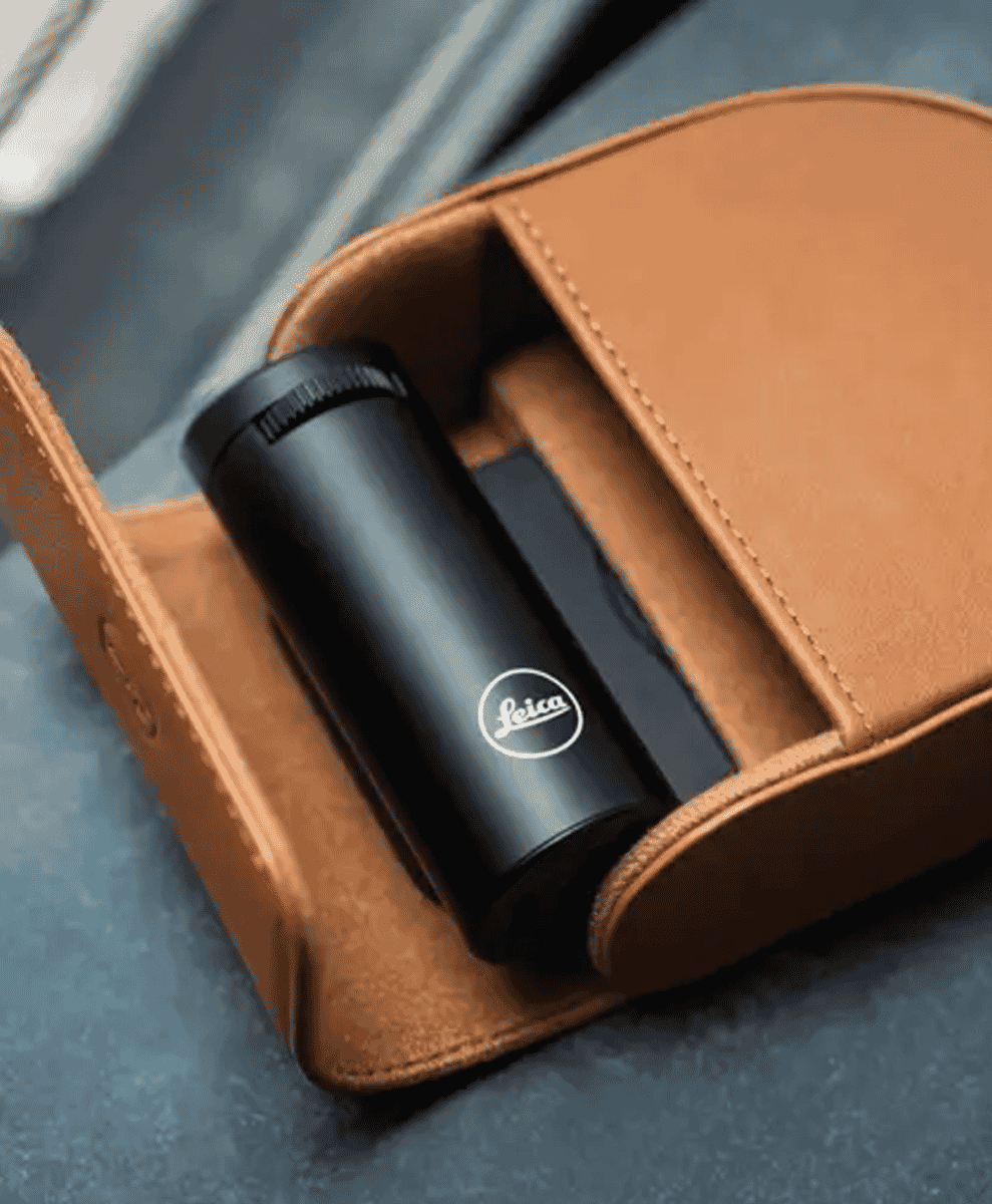 A black Leica cylindrical accessory resting in a premium tan leather case on a blue surface.