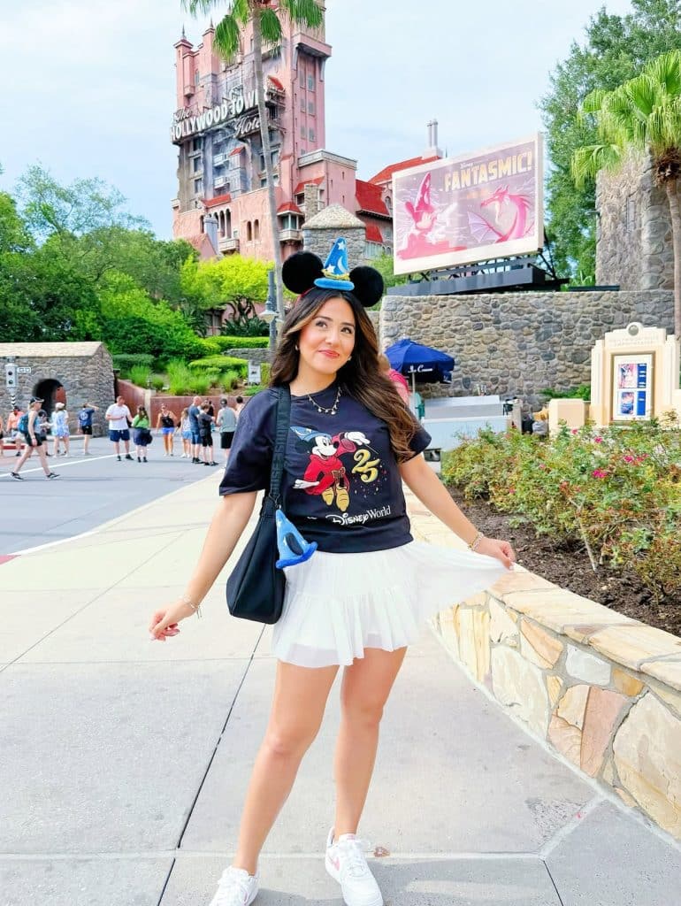 A woman in a black Mickey Mouse t-shirt and white pleated skirt with blue wizard ears in front of the Hollywood Tower Hotel.