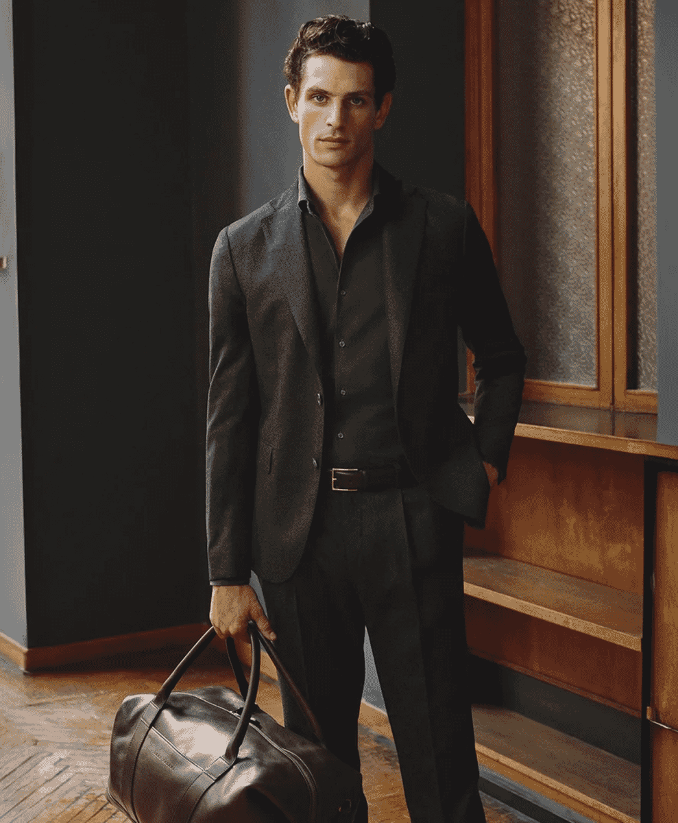 A man in a suit holding a leather duffel bag in a modern interior setting.