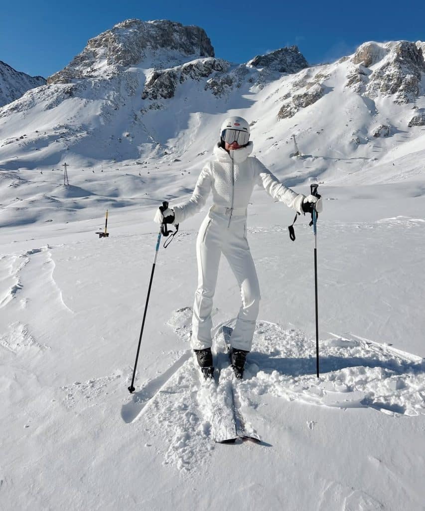 Skier in a full white ski suit and goggles with arms outstretched on a snowy slope.