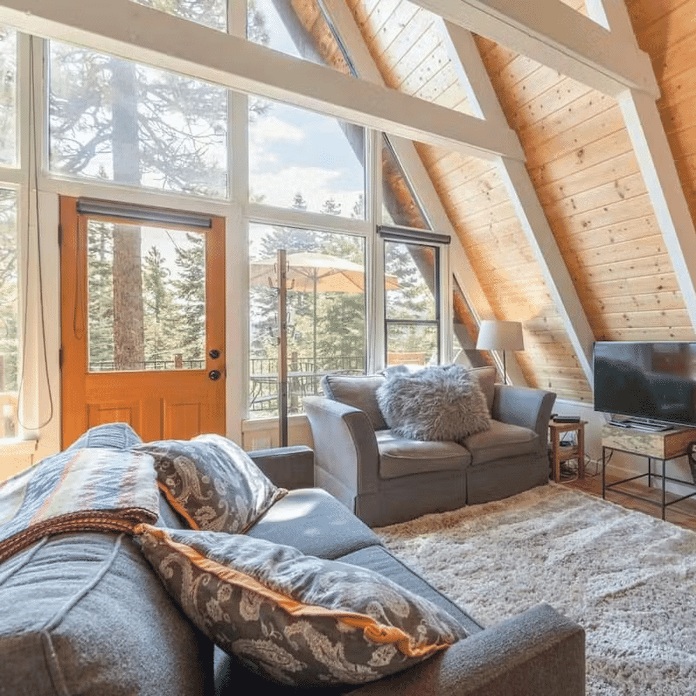 Cozy cabin living room with gray sofas and vaulted wooden ceiling looking out to a forest.