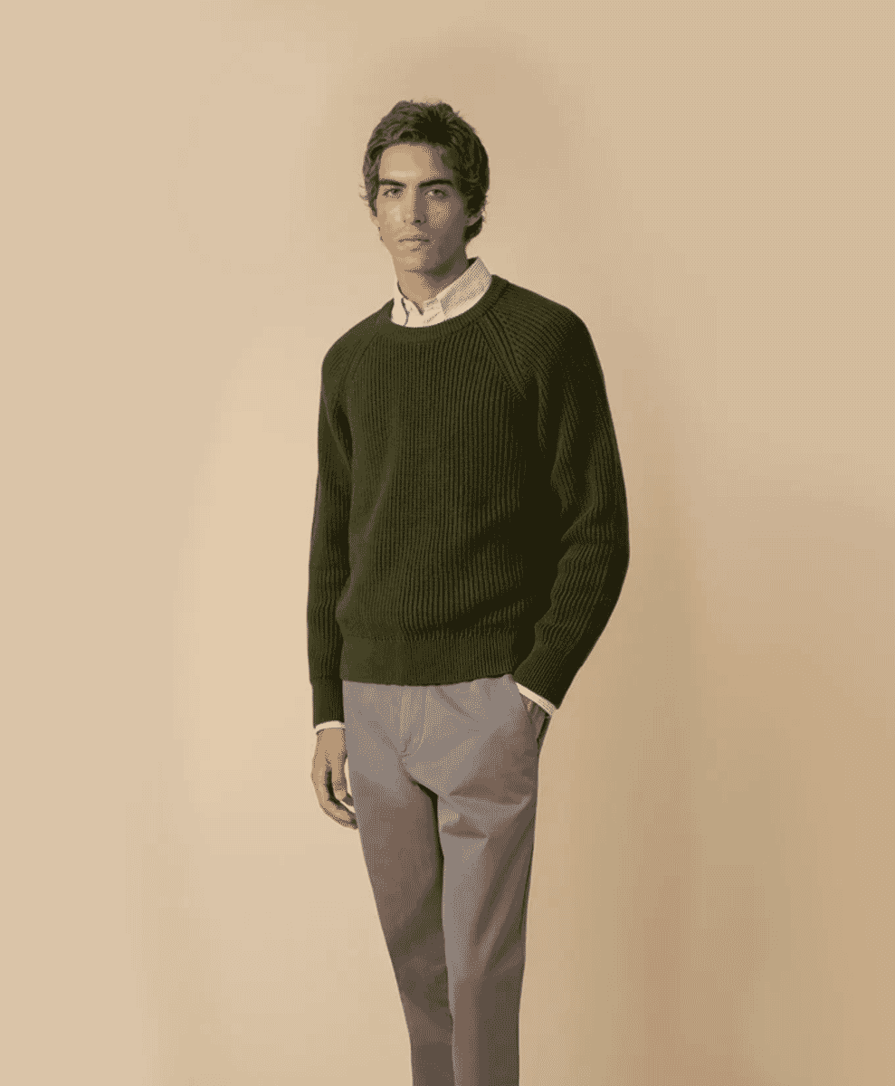 A man modeling a forest green ribbed knit sweater over a white dress shirt and grey trousers.