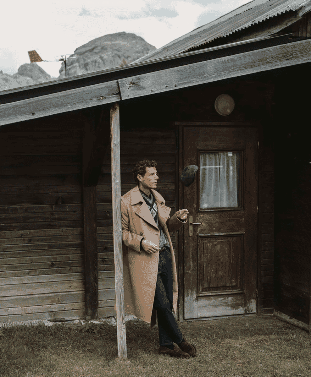 A man in a beige wool coat standing outside a rustic wooden mountain cabin.