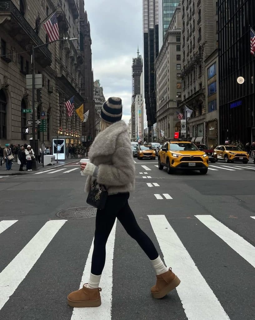 Woman crossing a city street in a fluffy grey coat, black leggings, and a striped beanie.