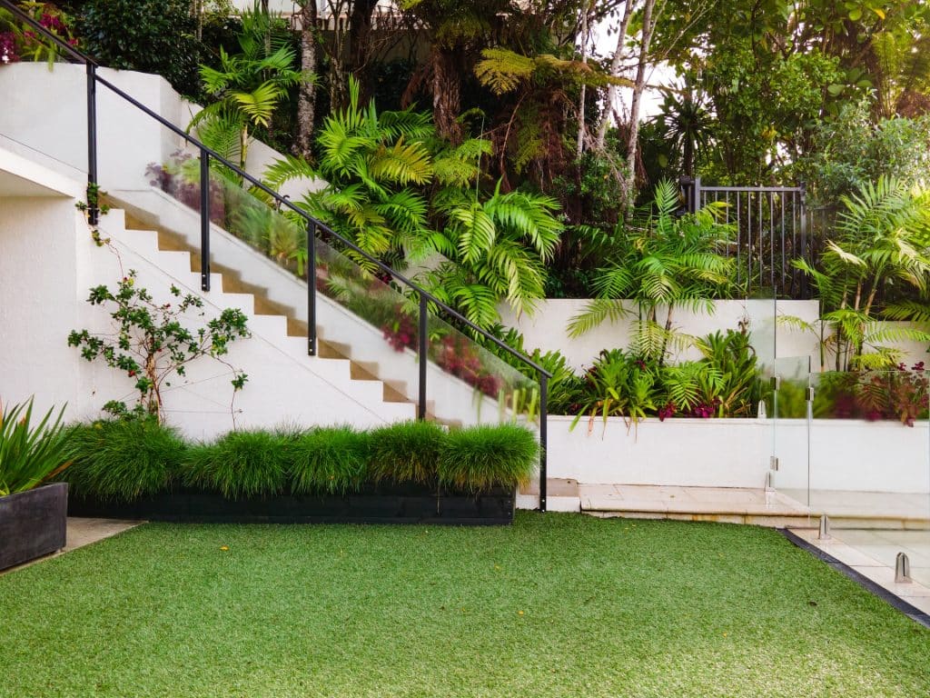 Modern garden design featuring white stairs with glass railings and tropical greenery.