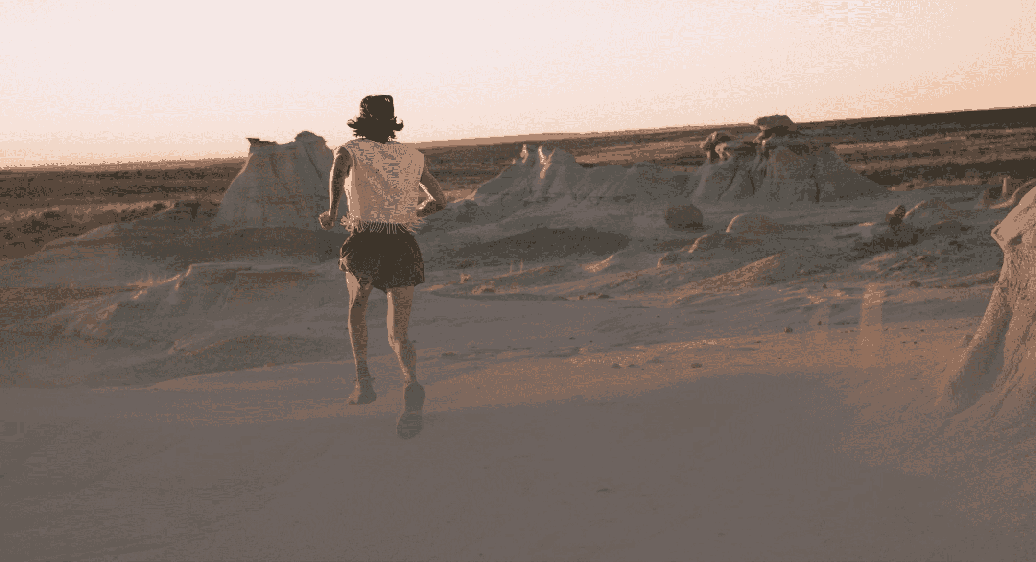 A person runs in a desert wearing a white fringed sleeveless top and athletic gear at sunset.