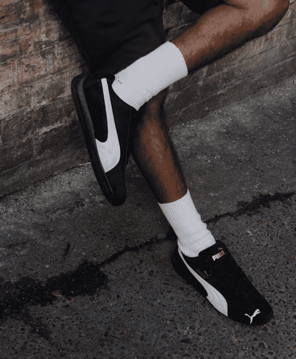 Person seated in black Puma Speedcat sneakers against a brick backdrop.