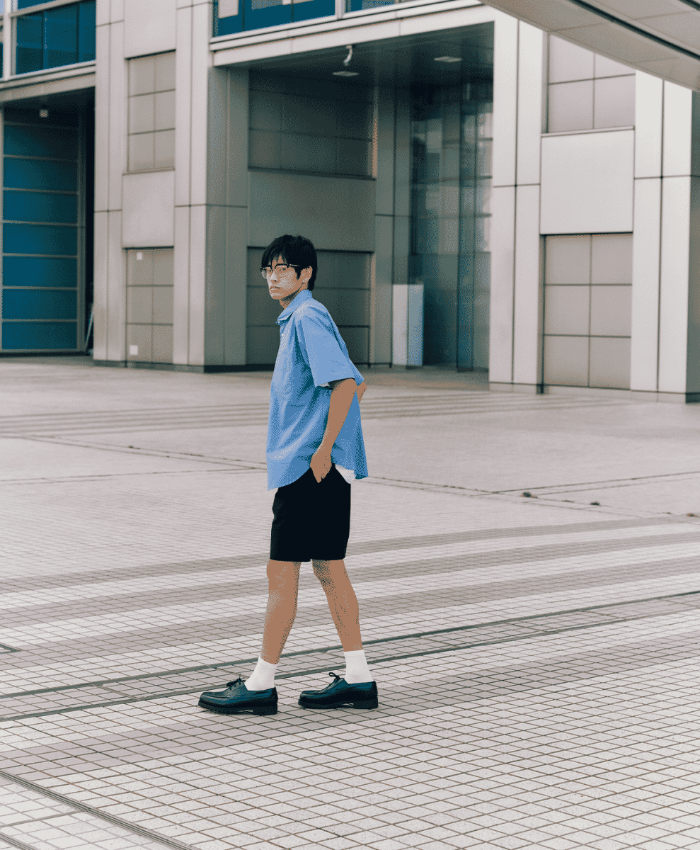 Man in an oversized light blue short-sleeve shirt and black shorts walking across a modern tiled plaza.