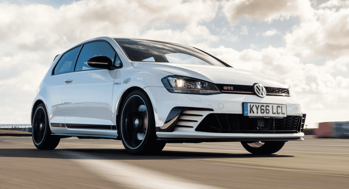 A white Volkswagen Golf GTI speeding across a racetrack under overcast skies.