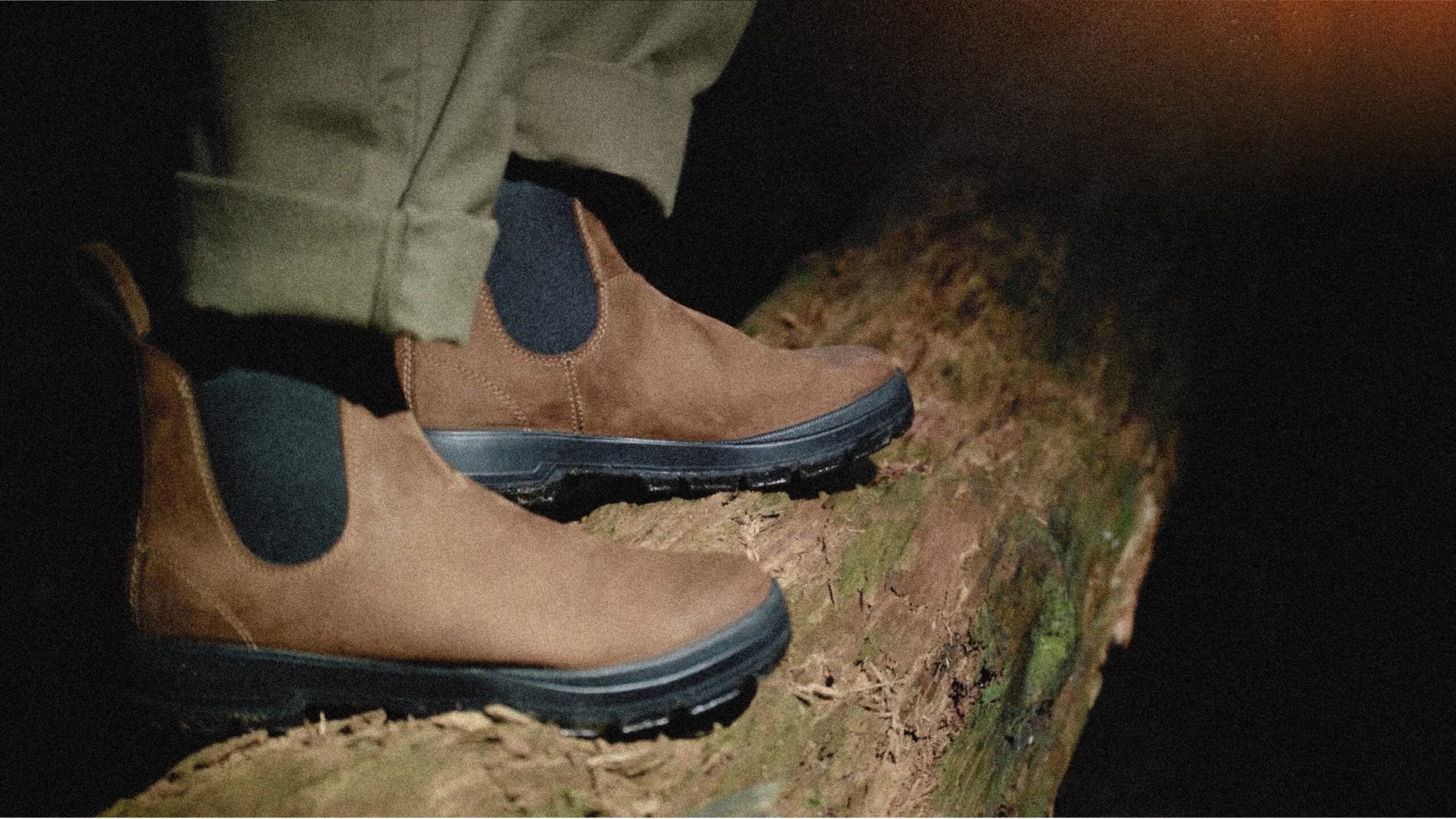A person wearing brown suede Chelsea boots and olive pants standing on a mossy log in the forest.