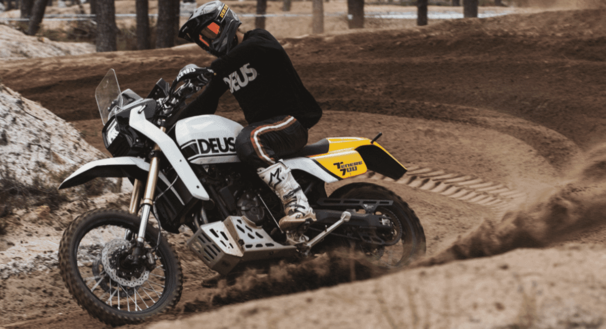 Action shot of the Deus dirt bike kicking up a cloud of dust on an off-road track.