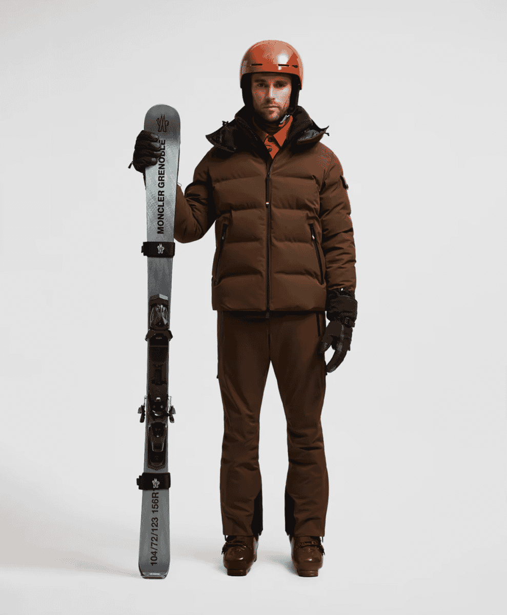A model in a brown puffer jacket and matching ski pants holding a Moncler Grenoble ski.