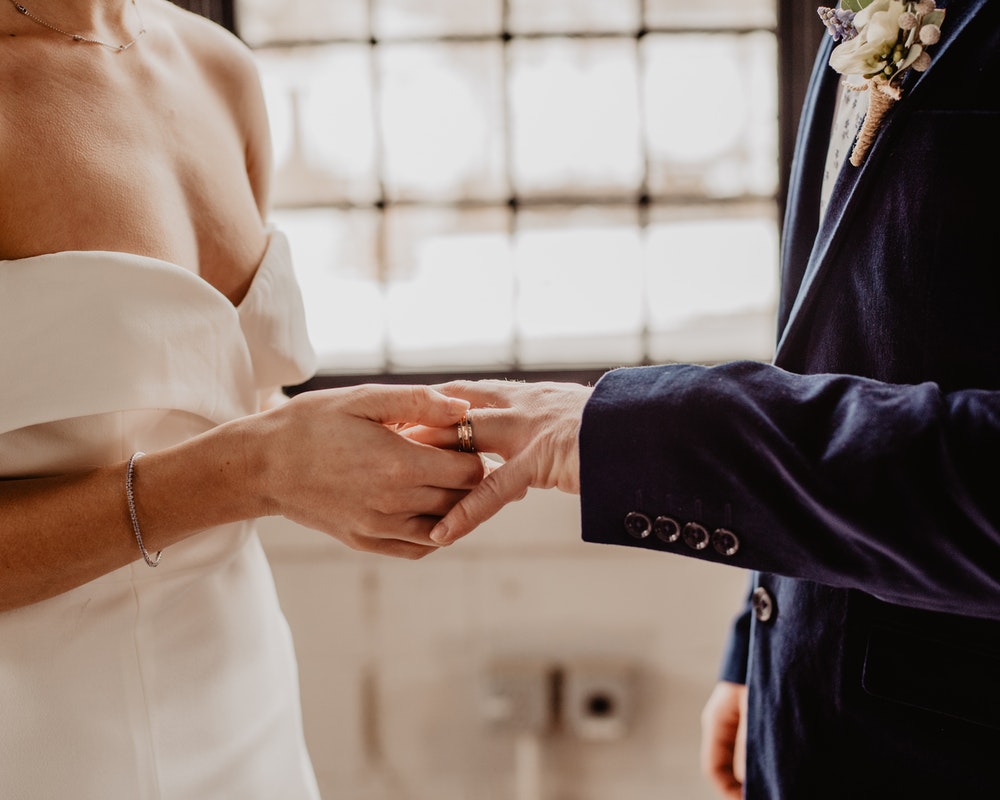 Groom in a navy suit receiving a wedding band during a ceremony with soft lighting.