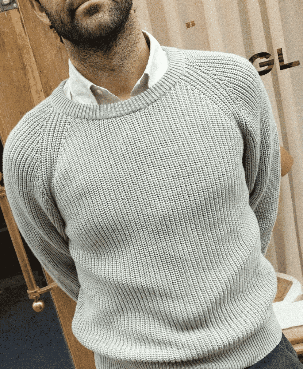 Man wearing a gray ribbed sweater over a collared shirt in a minimalist indoor setting