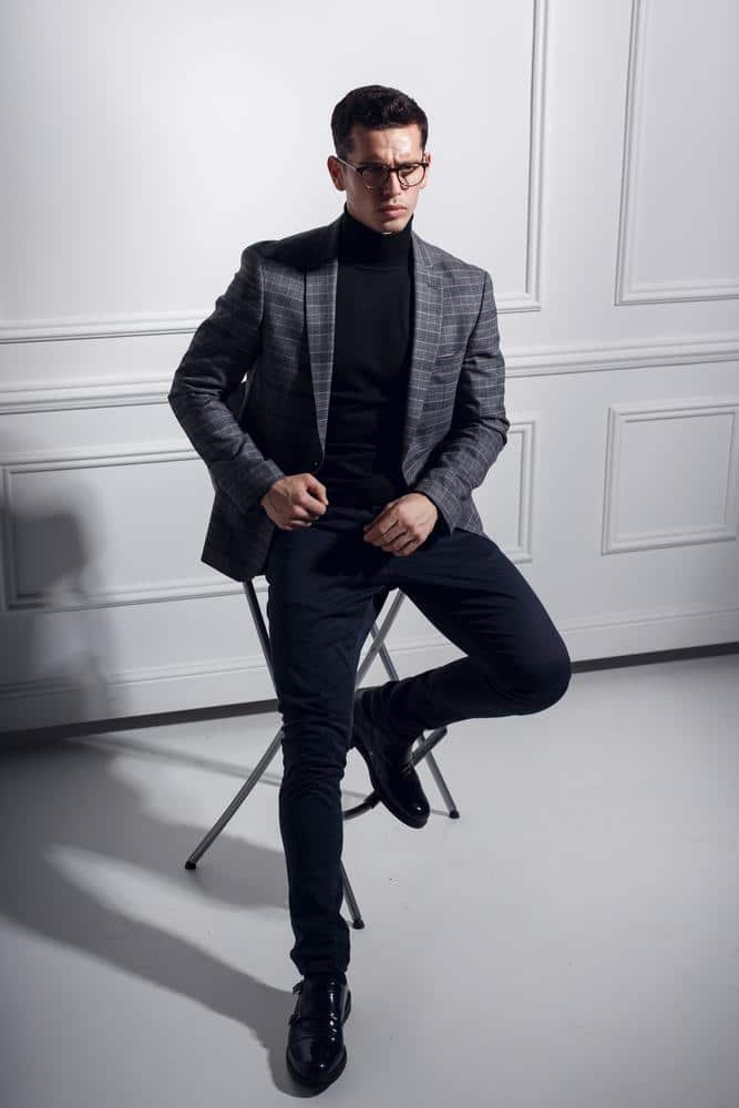Man in a grey plaid blazer and black turtleneck sweater posing in a studio setting.
