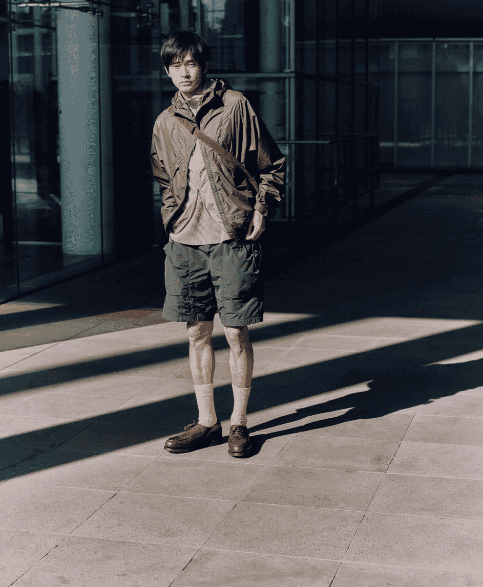 A man in a brown windbreaker and olive shorts paired with white socks and leather loafers in a sunny plaza.