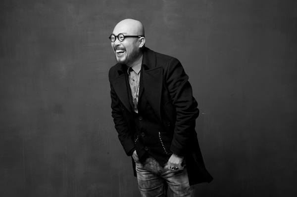 A black-and-white portrait of designer Lie Sang Bong wearing round glasses and a dark double-breasted coat, laughing against a gray backdrop.