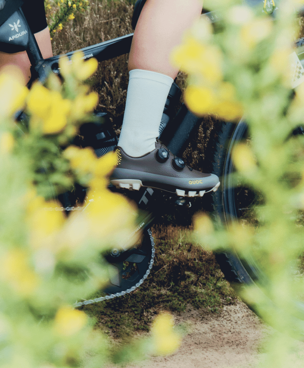 A cyclist pedaling through a vibrant field of yellow wild flowers wearing QUOC mountain bike shoes.
