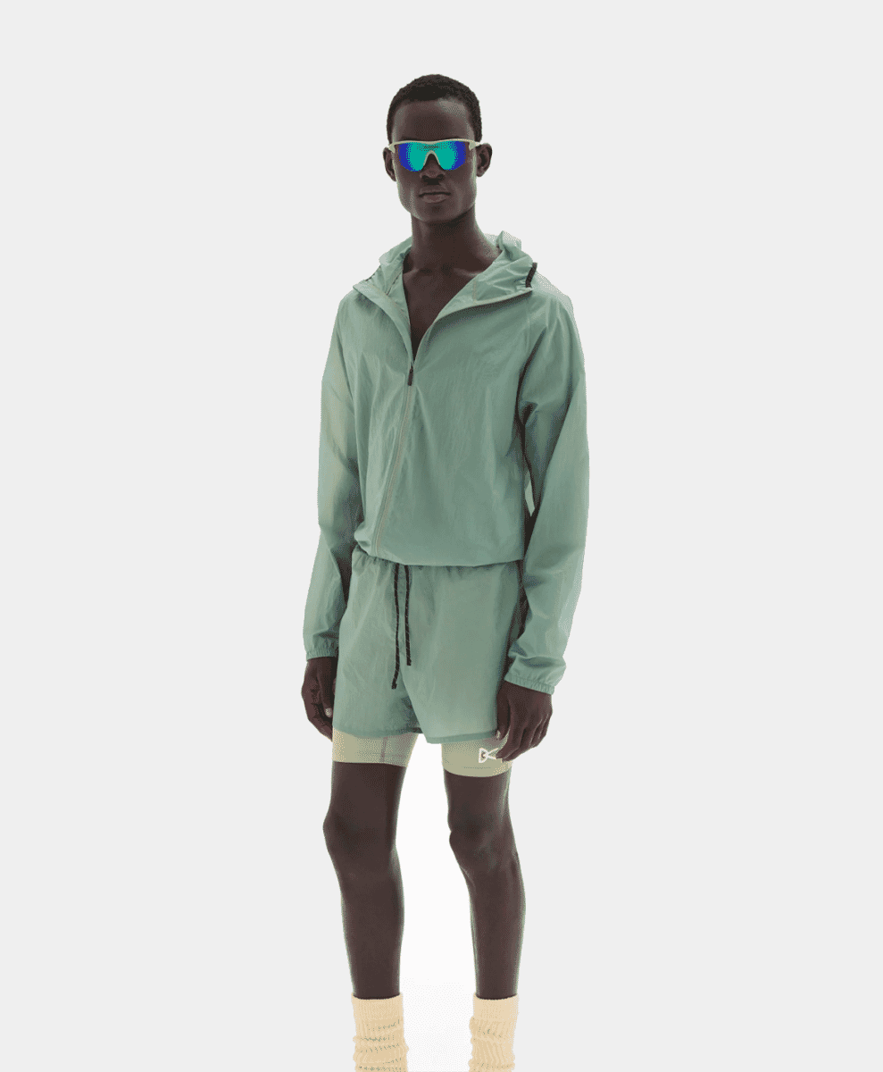 Model in a mint-green hooded windbreaker and reflective sunglasses against a white background.