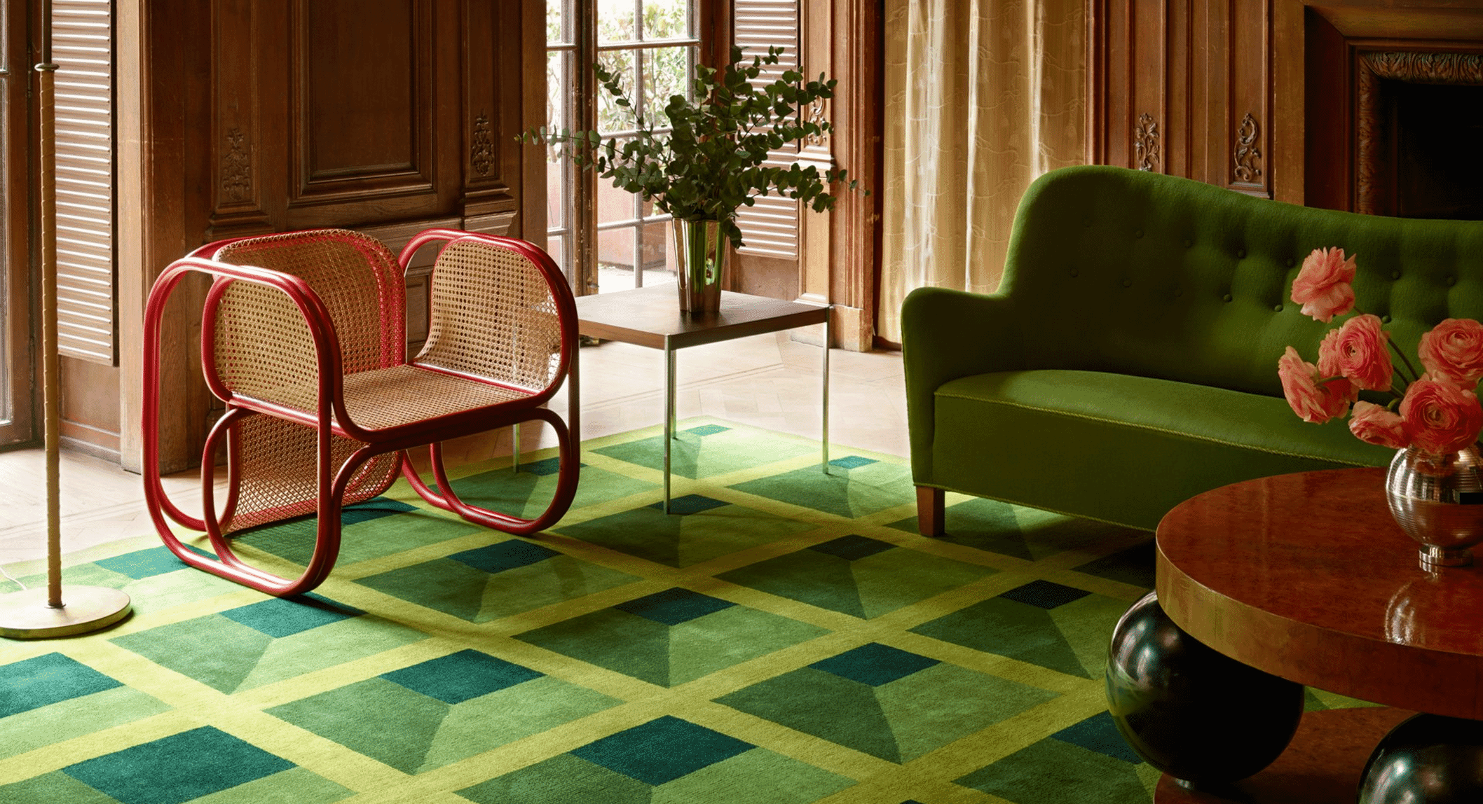A living room with a green tufted sofa, red cane chairs, and a geometric green rug against wooden paneling.