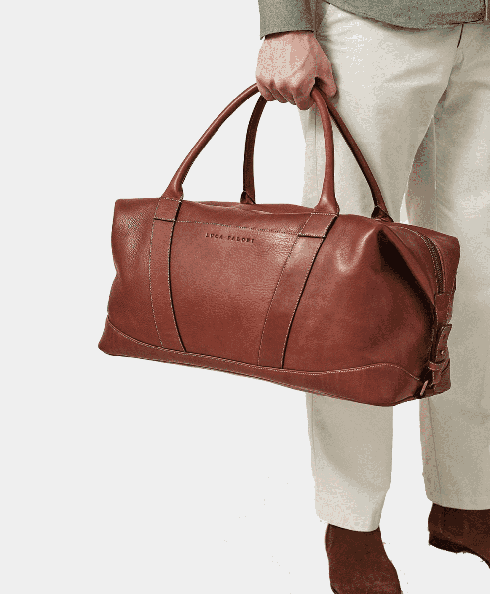 Man in a light gray sweater and cream trousers carrying a brown leather duffel bag.