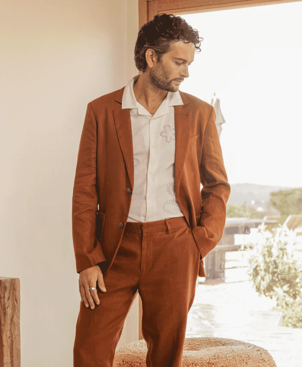 Man in a coordinated brown suit with a white embroidered shirt standing near a bright window.