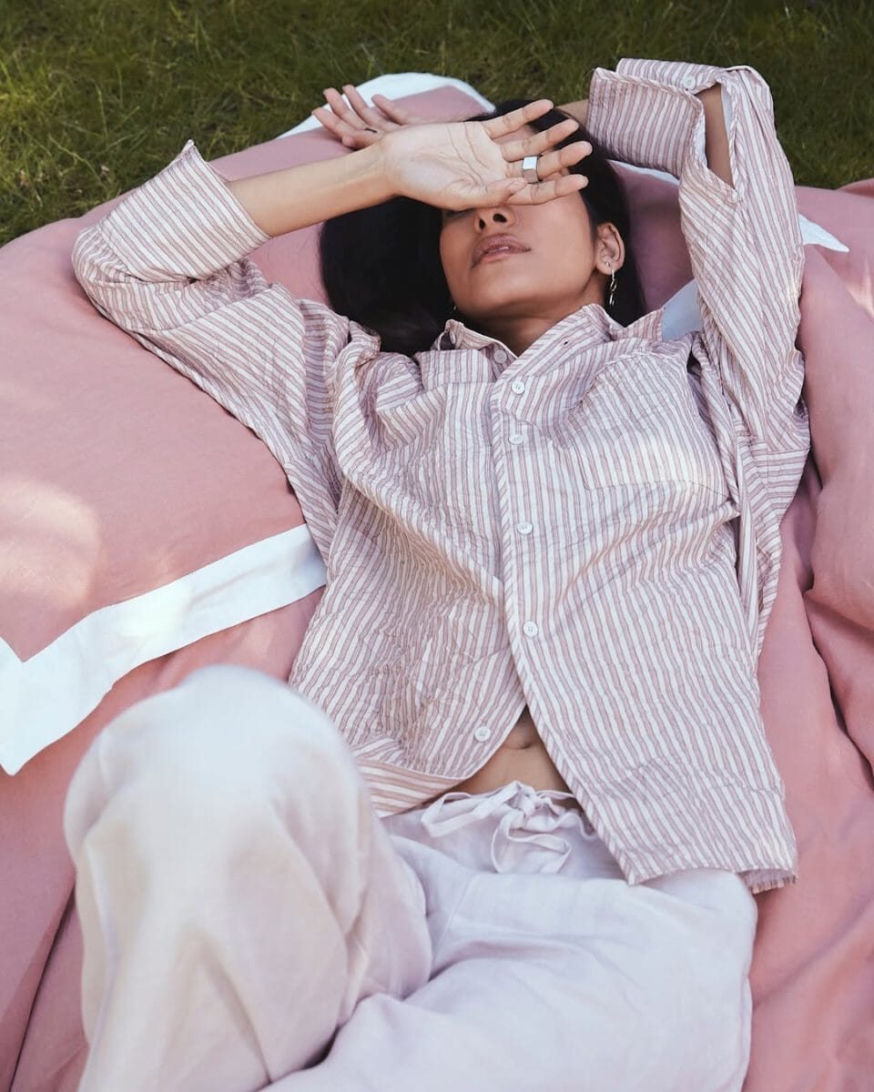 A woman in a pink and white striped shirt and light pants lies on a blanket outdoors, shielding her eyes from the sun.