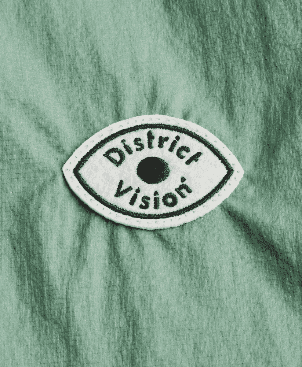 Close-up of a District Vision eye-shaped patch on green technical fabric.