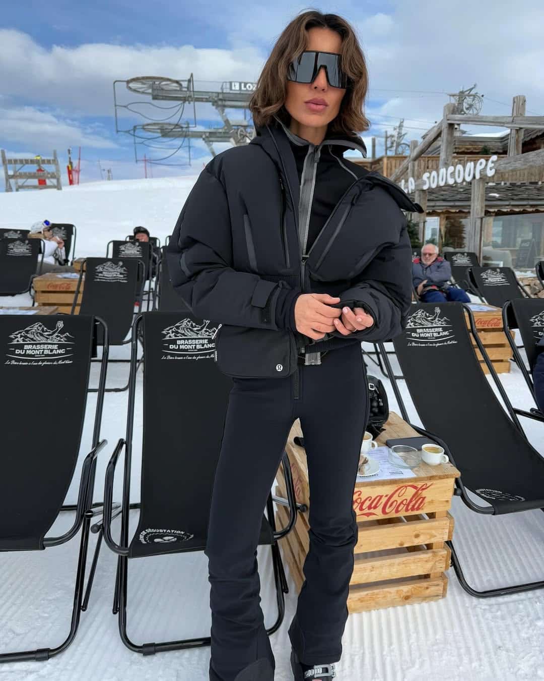 Woman in a black puffer jacket and oversized sunglasses on a snowy mountain terrace.