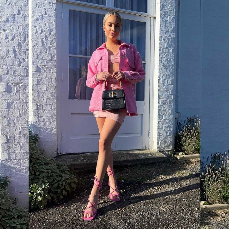 A woman in a vibrant pink mini-skirt set and strappy high heels posing against a white brick wall.