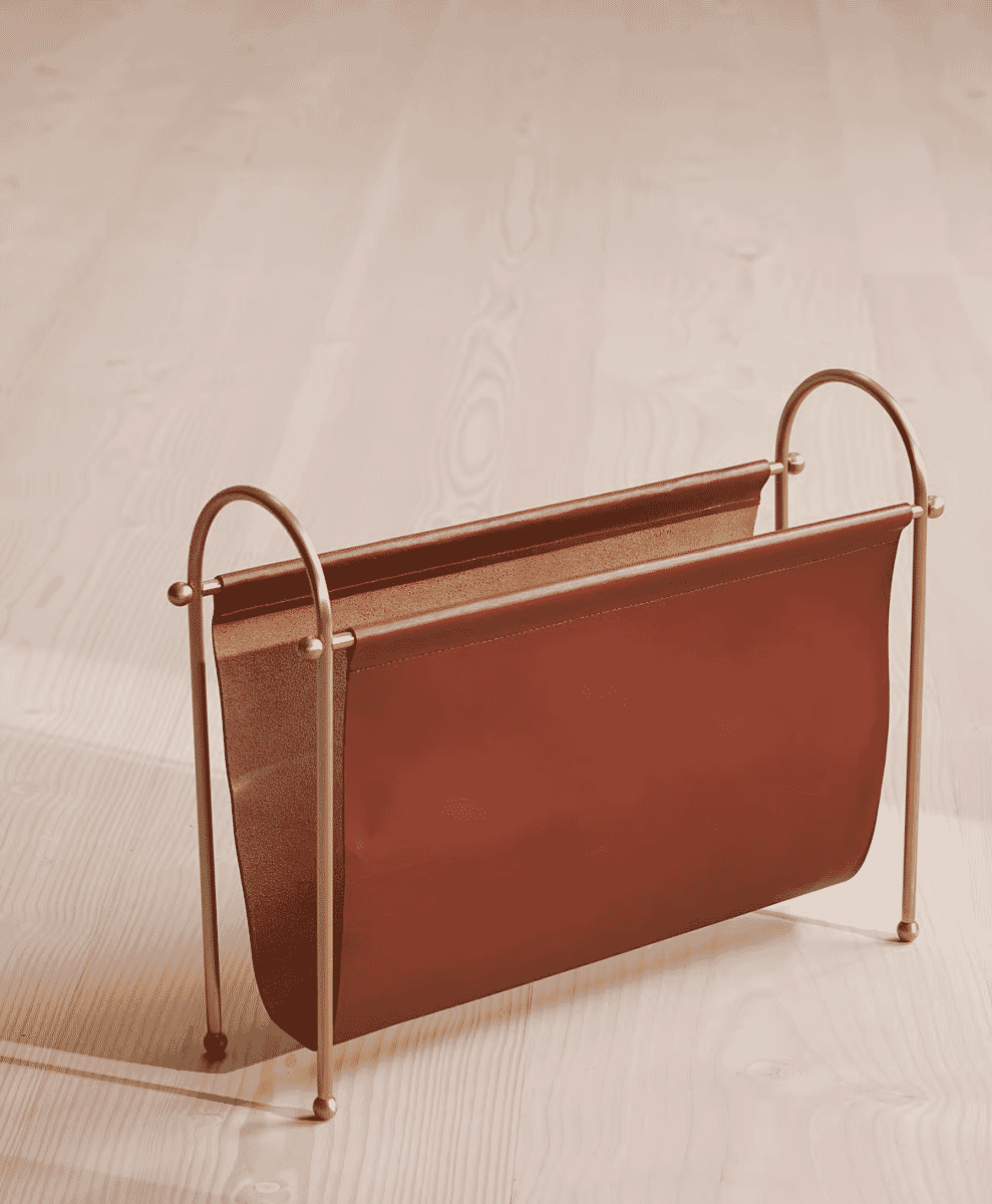 A minimalist magazine rack with a brass frame and brown leather insert on a wooden surface.