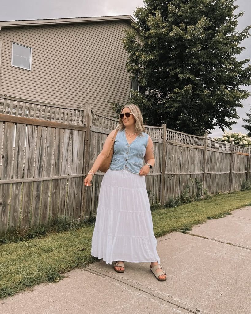 A woman in a light blue denim vest and white tiered maxi skirt on a sidewalk.