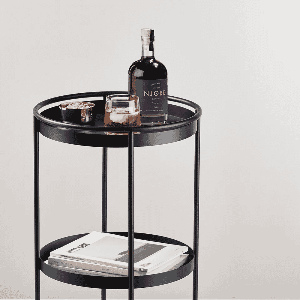 A minimalist black metal two-tier side table holding a gin bottle, glassware, and design magazines.