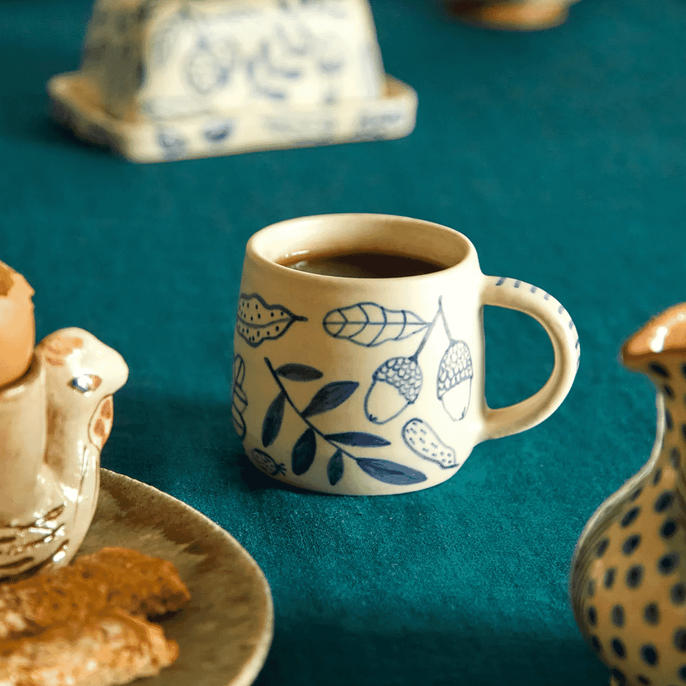 A hand-painted ceramic mug with blue botanical illustrations on a teal tablecloth.