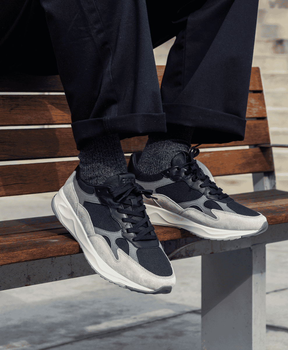 A person on a wooden bench wearing cuffed trousers and textured grey sneakers.