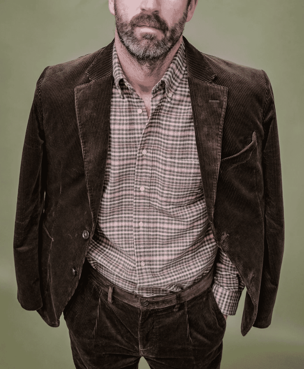 A man wearing a brown corduroy blazer over a checkered button-up shirt.