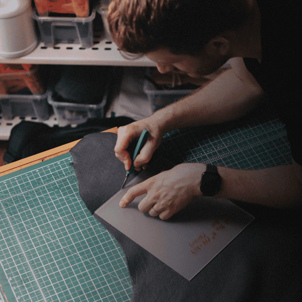 An artisan meticulously cutting tan leather with a precision knife on a green gridded mat.