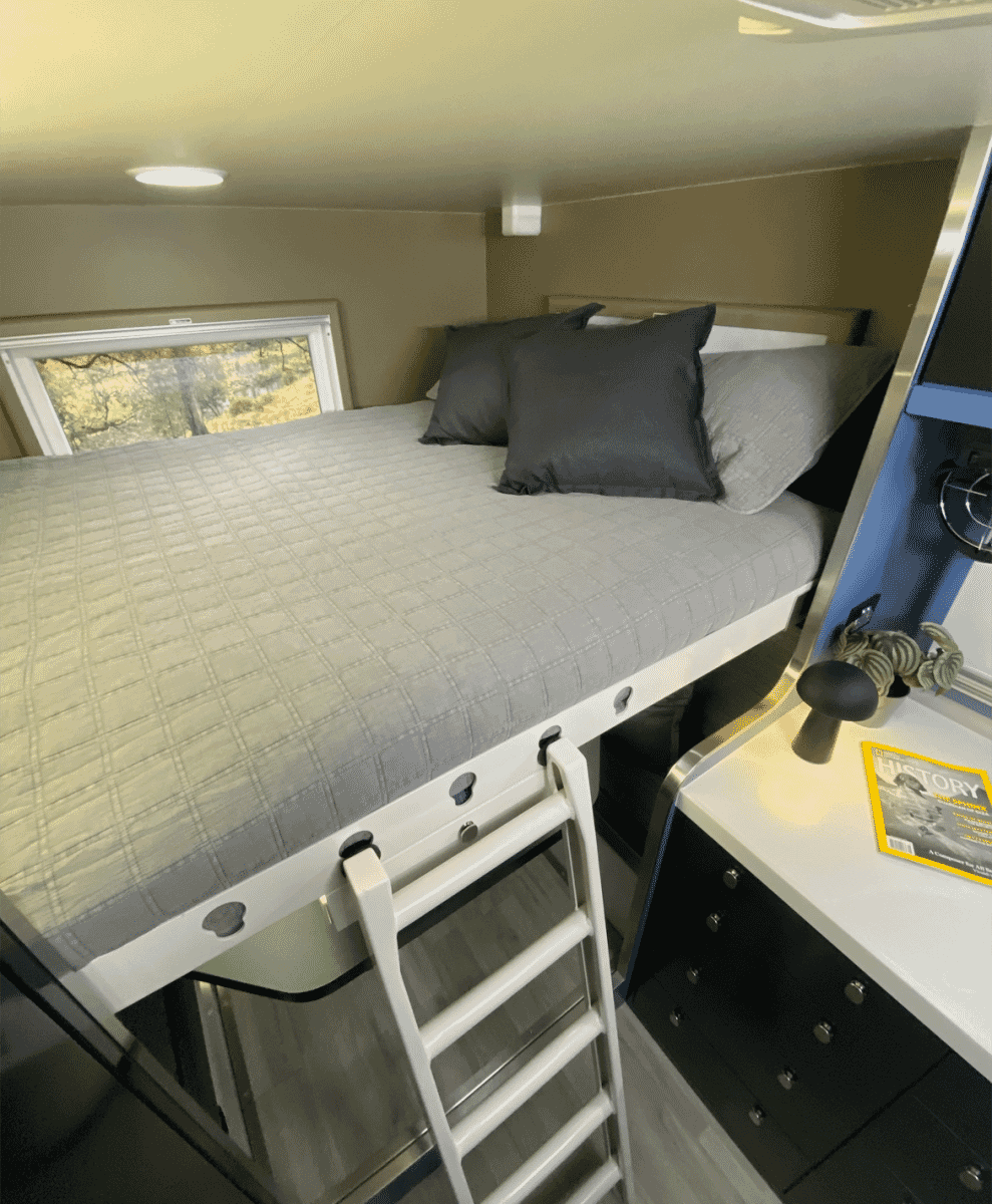 Modern RV interior showing a gray loft bed, wooden cabinetry, and a clean ladder system.