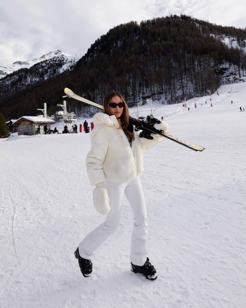 A woman in a chic all-white puffer jacket and matching pants carrying skis on a slope.