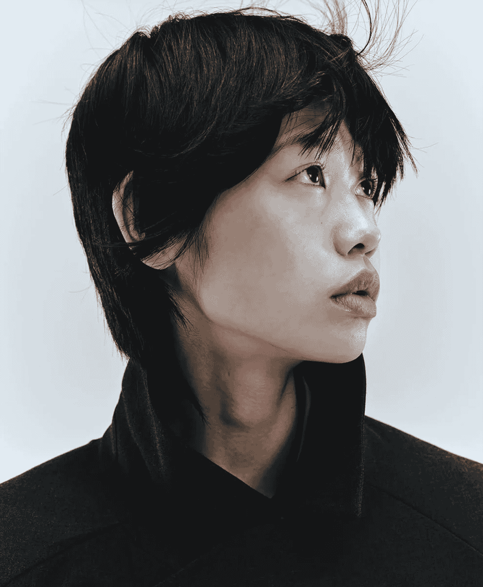 A close-up studio portrait of a person wearing a high-collared minimalist black coat.