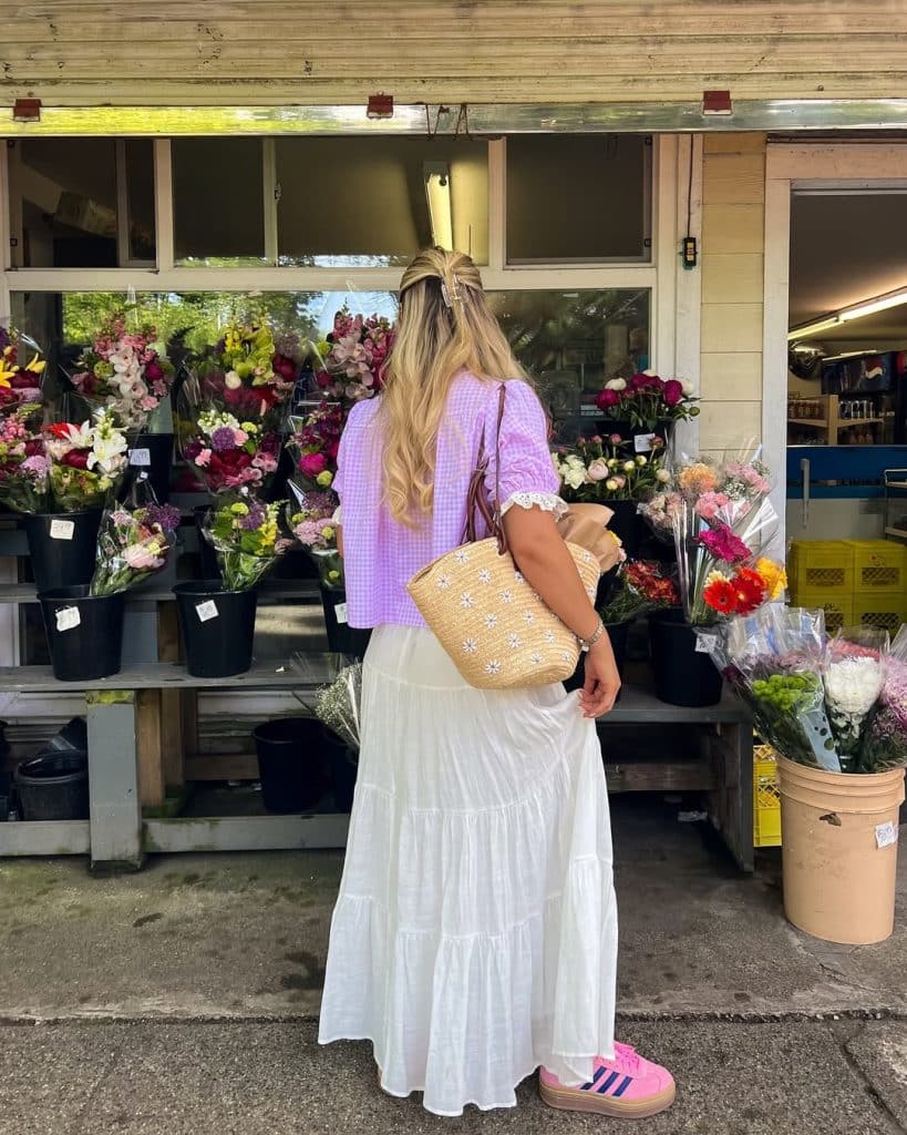A woman in a lavender gingham top and white tiered skirt at an outdoor flower market.
