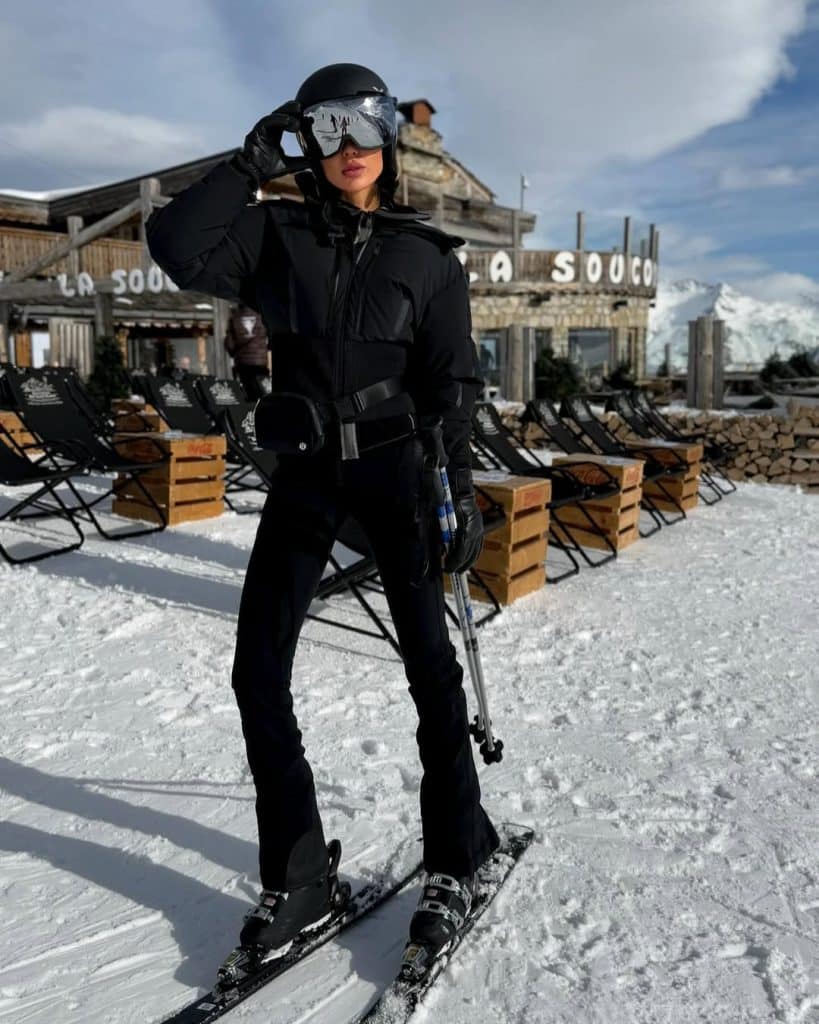 A woman in all-black insulated ski attire with goggles and poles at a mountain lodge.