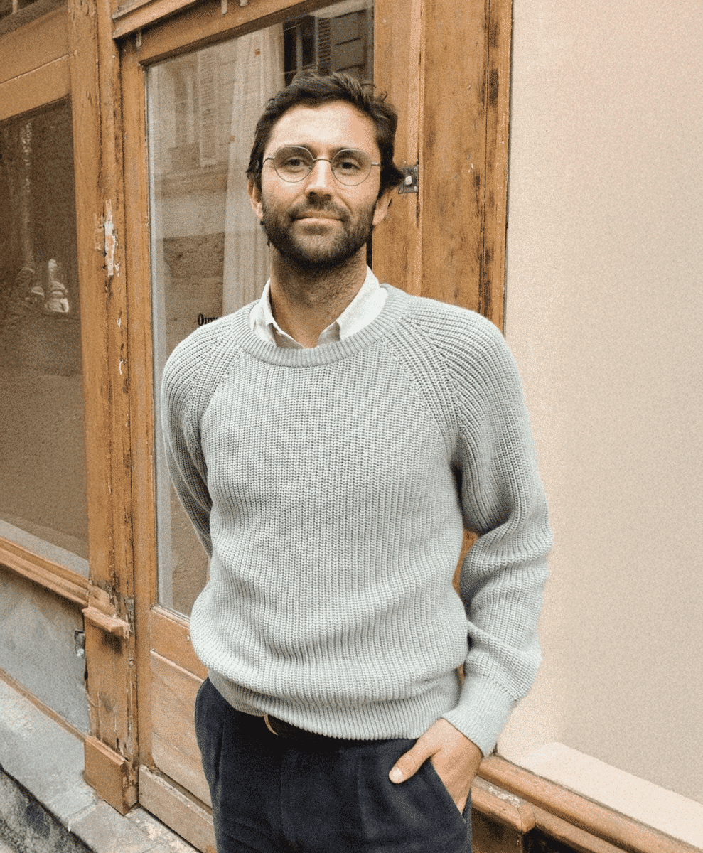 Man in a light gray ribbed knit sweater and dark trousers leaning against a wooden door