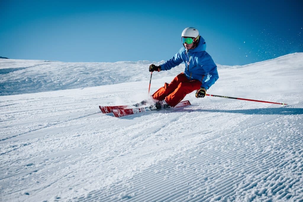 A skier in a blue jacket and red pants carving through fresh snow on a sunny mountain slope.