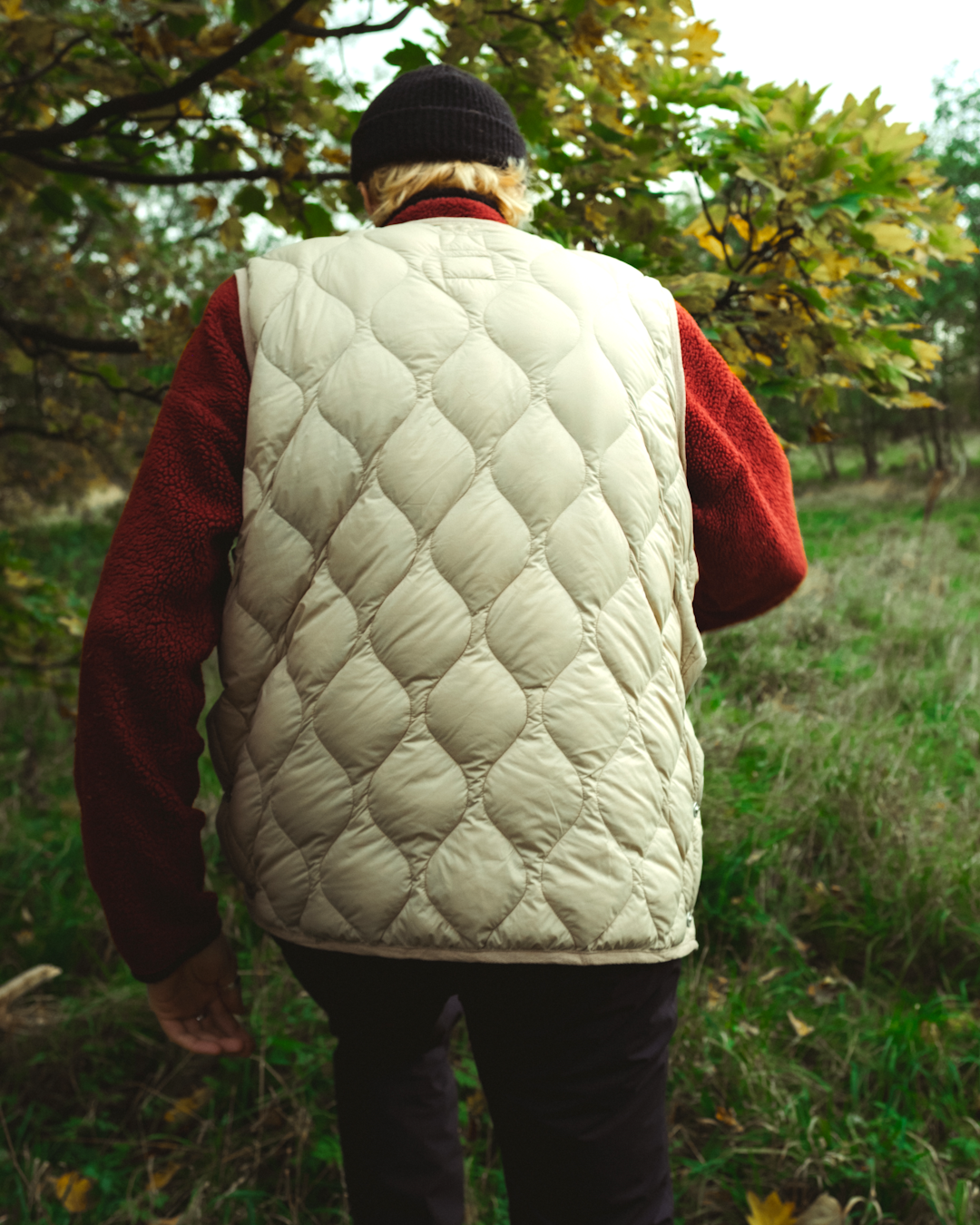 A person viewed from behind wearing a cream diamond-quilted puffer vest in a wooded path.