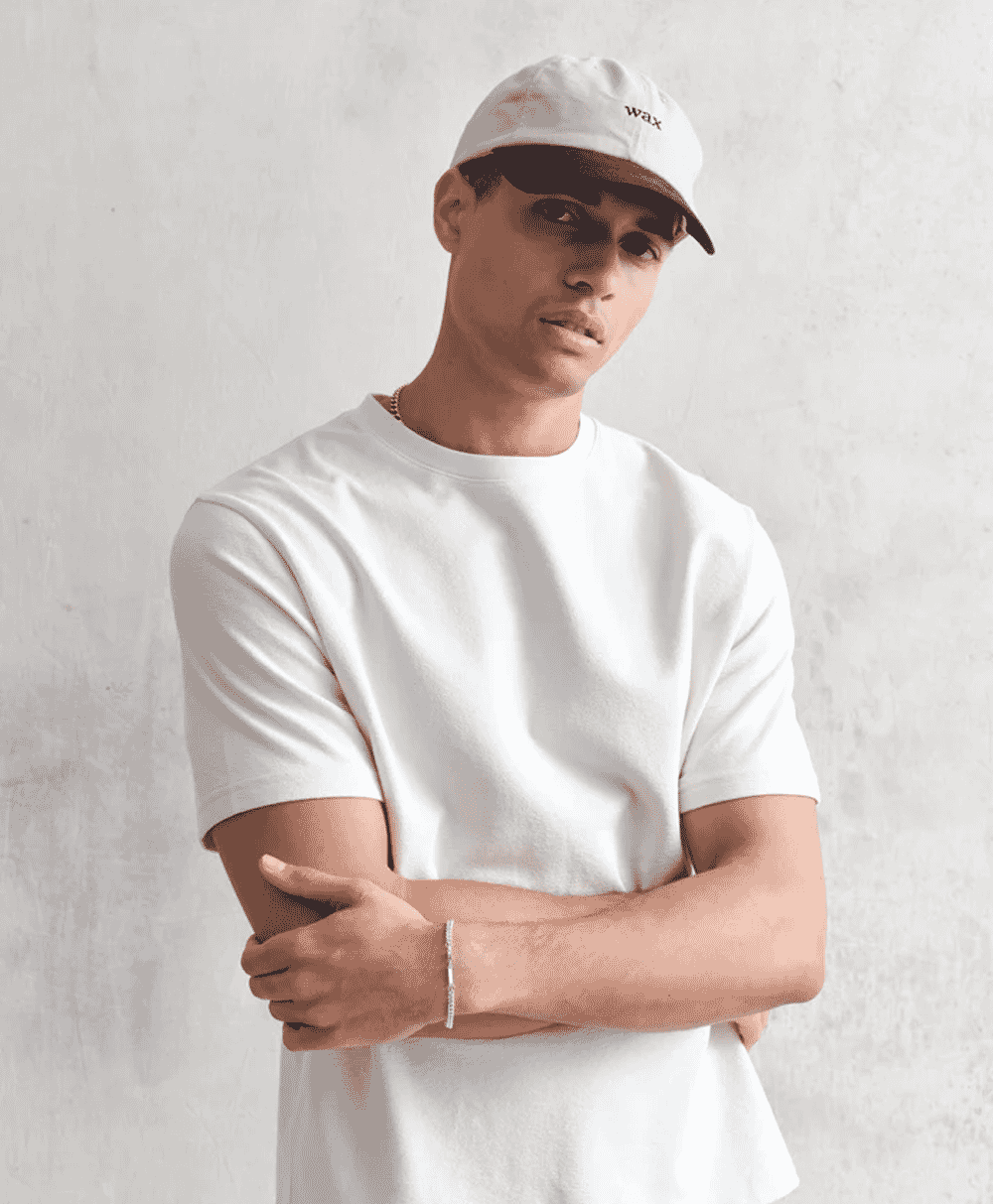 Man in a white 'wax' embroidered cap against a white wall.