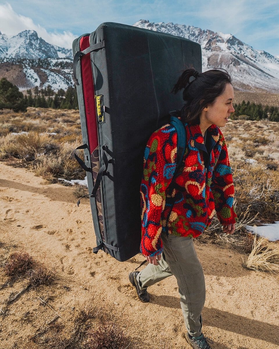 Woman in a colorful patterned fleece hiking through snow with a climbing crash pad.