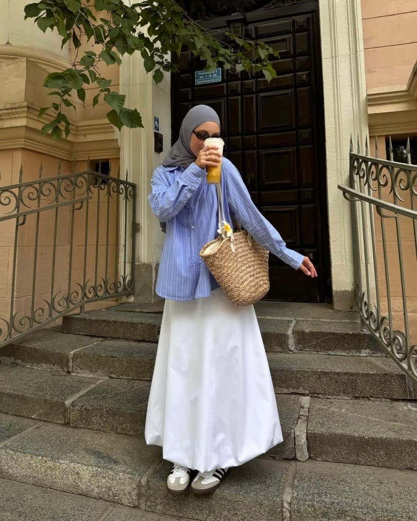 A woman in a blue striped shirt, white maxi skirt, and grey hijab on stone steps.